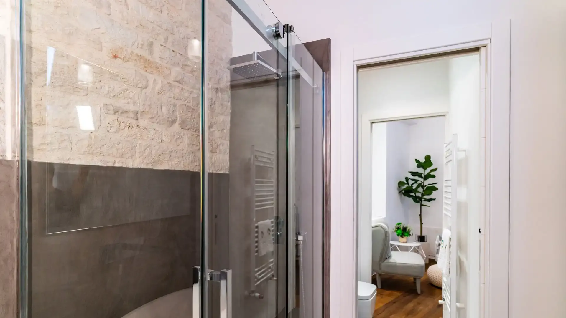 Salle de bain moderne avec douche en verre, murs en pierre et vue sur une pièce adjacente.