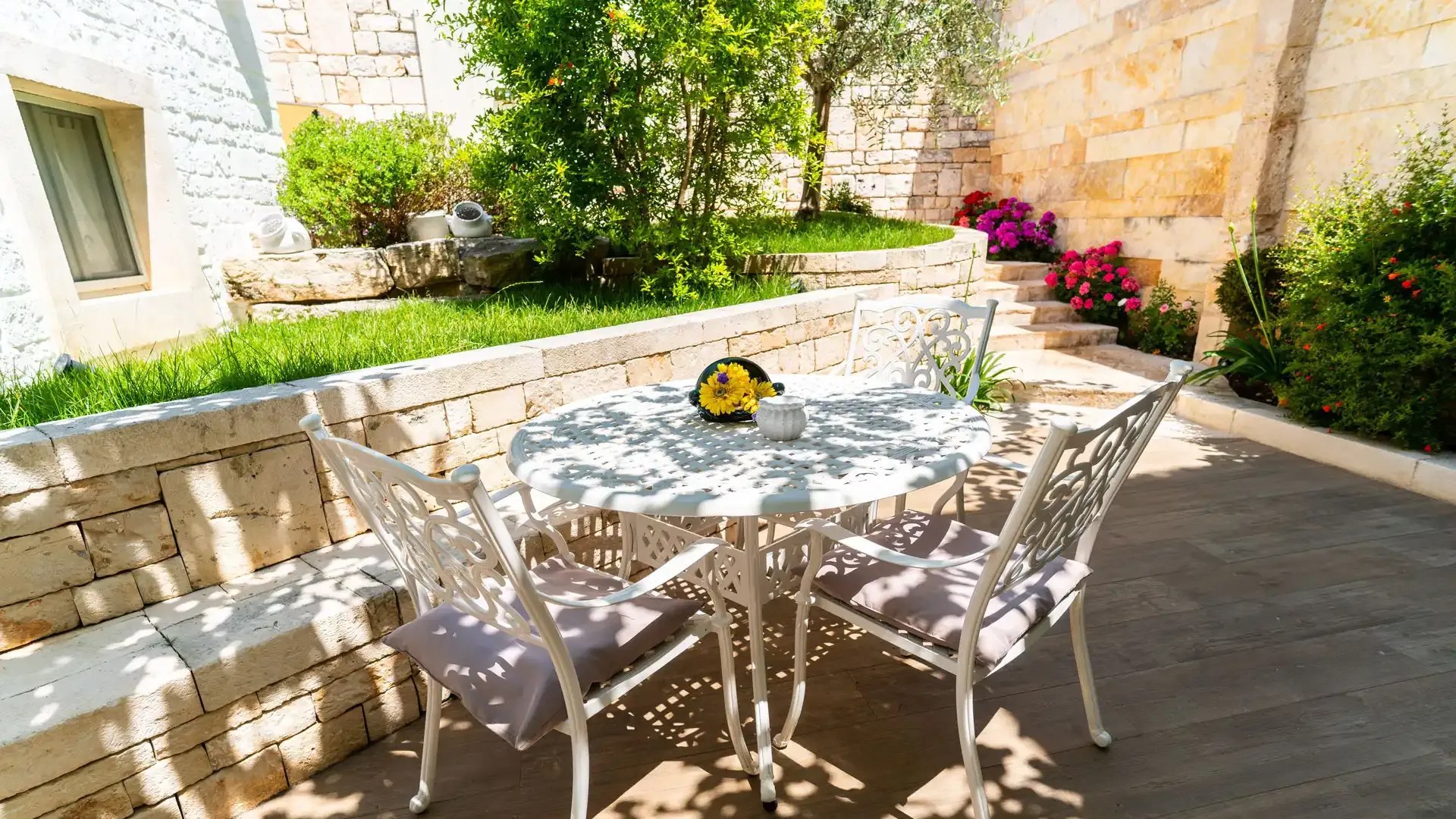 Jardin avec table et chaises en fer forgé, entouré de fleurs et de plantes.