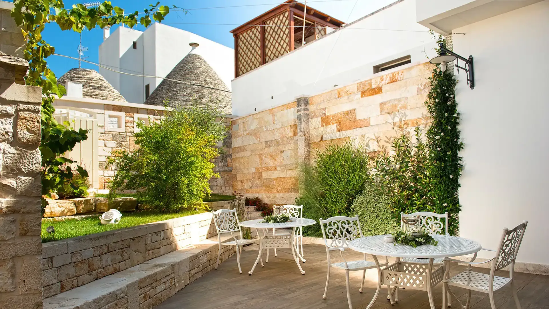 Jardin avec tables et chaises blanches, plantes et murs en pierre.